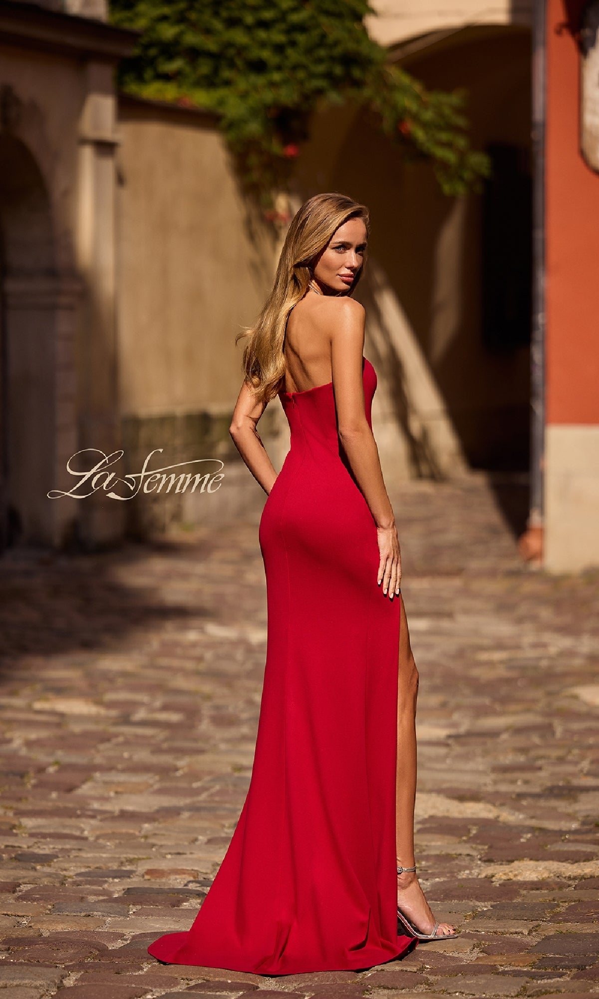 A woman in the Classic Strapless Long Prom Dress: La Femme 32784, featuring a thigh-high slit, stands on a cobblestone street and looks over her shoulder.